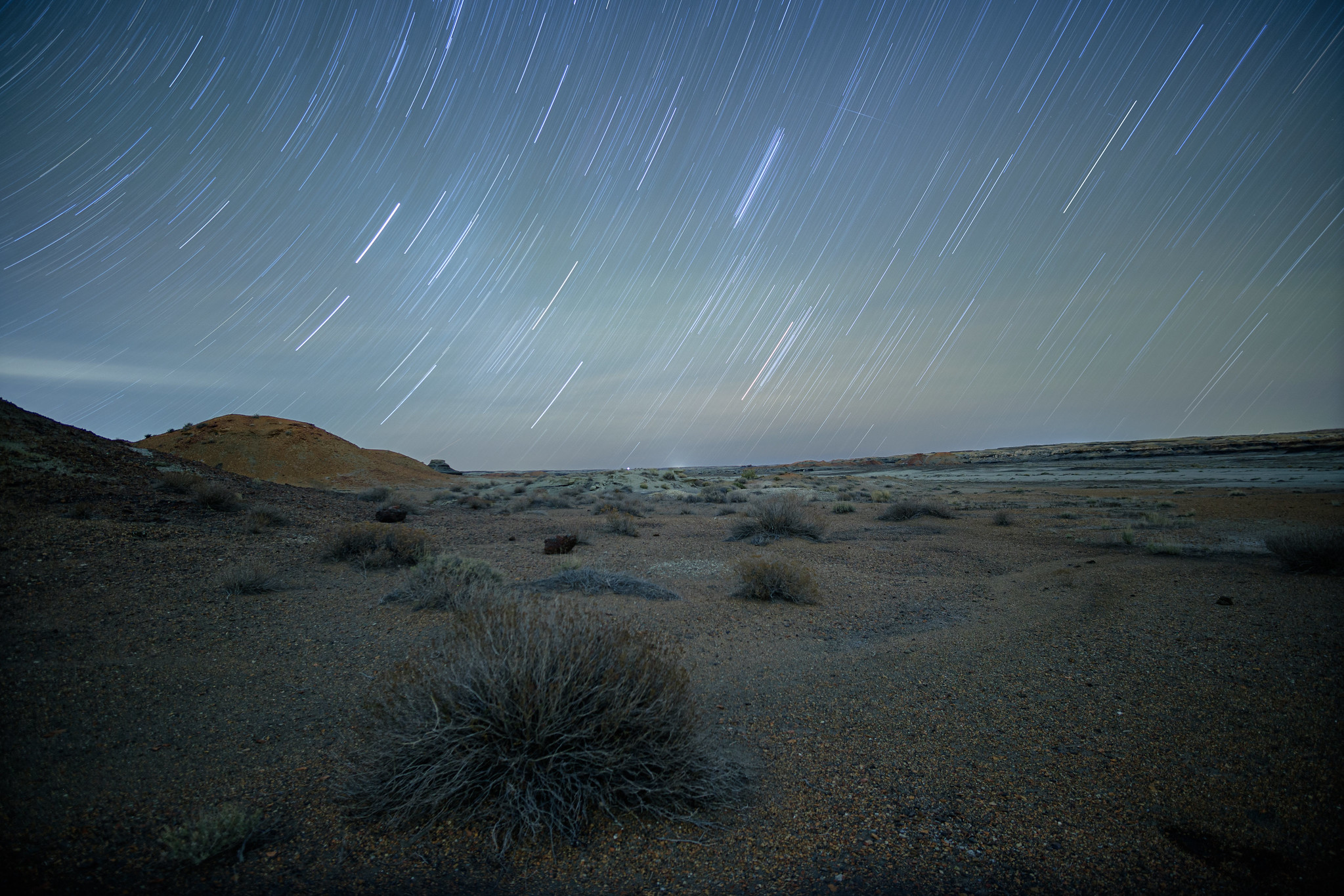 A landscape of a badlands in the Bisti/De-Na-Zin Wilderness. It is a long exposure image taken at night, and the stars are leaving long trails in the night sky.