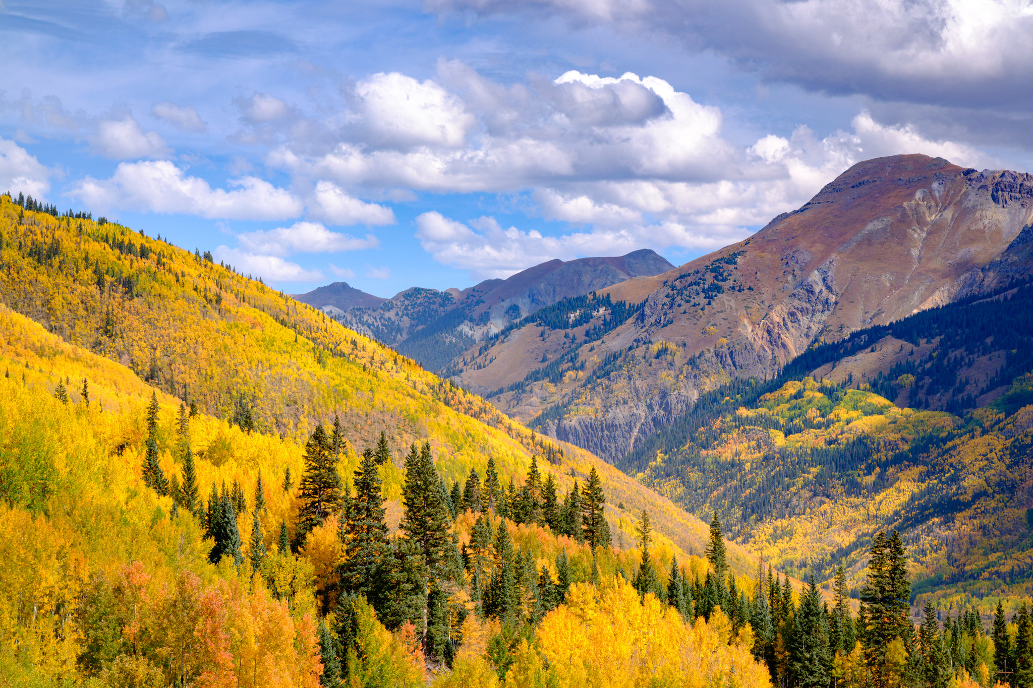 A landscape with yellow aspecs in the left half and reddish mountains in the right, with blue skies with mostly white clouds.